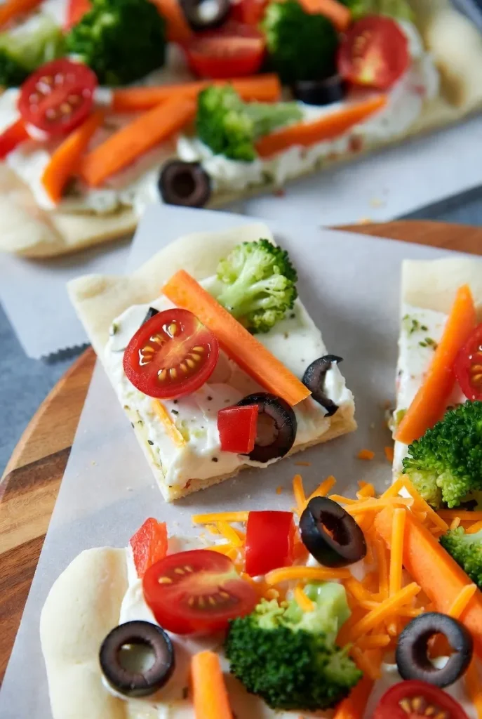 Close-up of vibrant veggie pizza squares featuring broccoli, tomatoes, carrots, olives, and cheese on a creamy base.