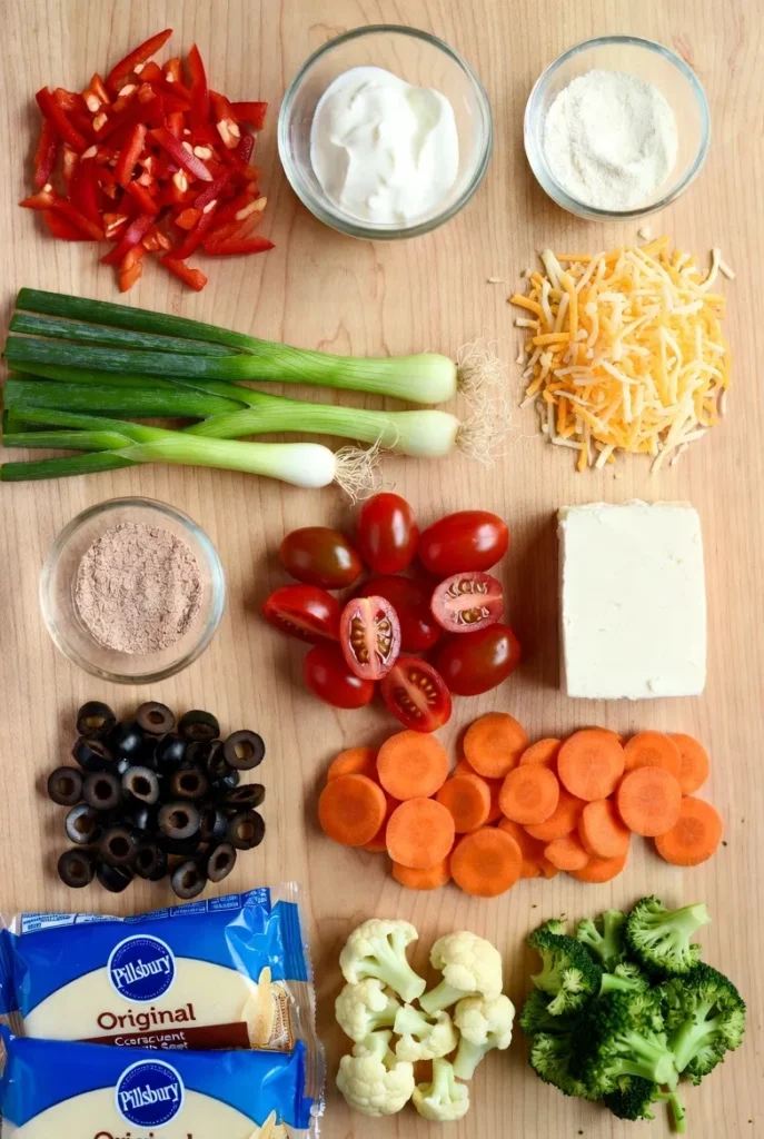 A colorful overhead flat lay of chopped vegetables and ingredients, including red bell peppers, cauliflower, broccoli, carrots, cherry tomatoes, olives, cream cheese, shredded cheddar, green onions, crescent dough, and bowls of sour cream and spices on a light wooden cutting board.