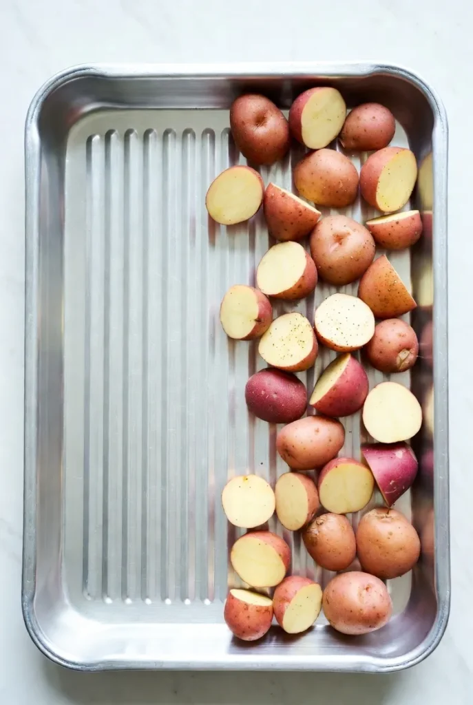 An overhead view of a silver baking sheet with scattered red and yellow potato wedges, lightly seasoned.