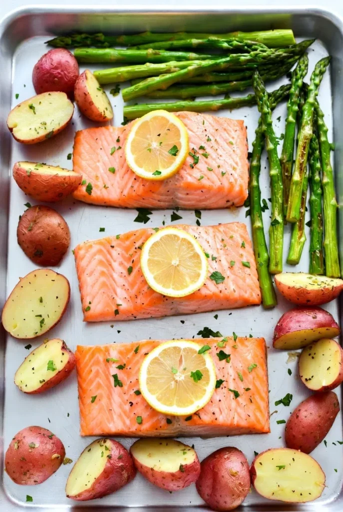 A full sheet pan of baked salmon with lemon, roasted asparagus, and red potatoes, viewed from above.