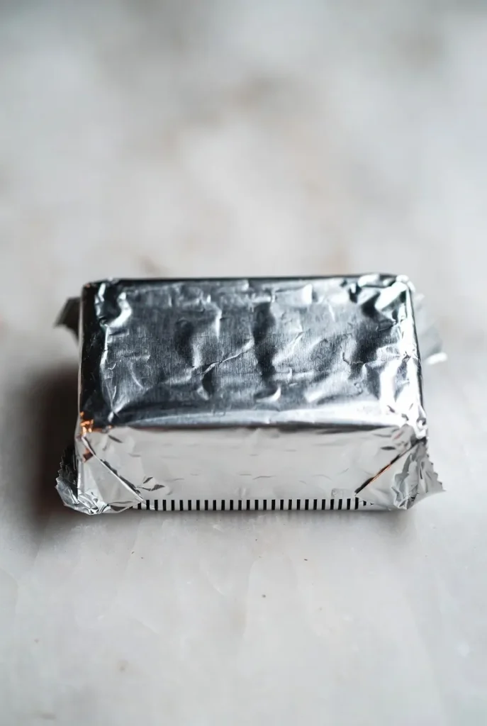 A silver foil-wrapped block of butter or cream cheese on a light marbled surface.
